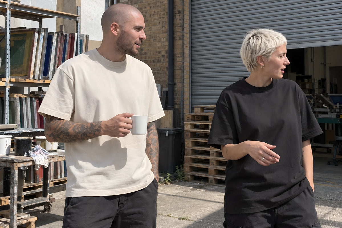 A man in his late twenties with a shaved head and a full sleeve of tattoos, in the bone ENVY Classic Tee, standing in the yard behind a screen-print shop talking to a woman with bleached cropped hair in a black oversized tee, both half turned away from each other mid-conversation, a roller shutter and stacked pallets behind them. Daytime, hard sun, no candles lit.