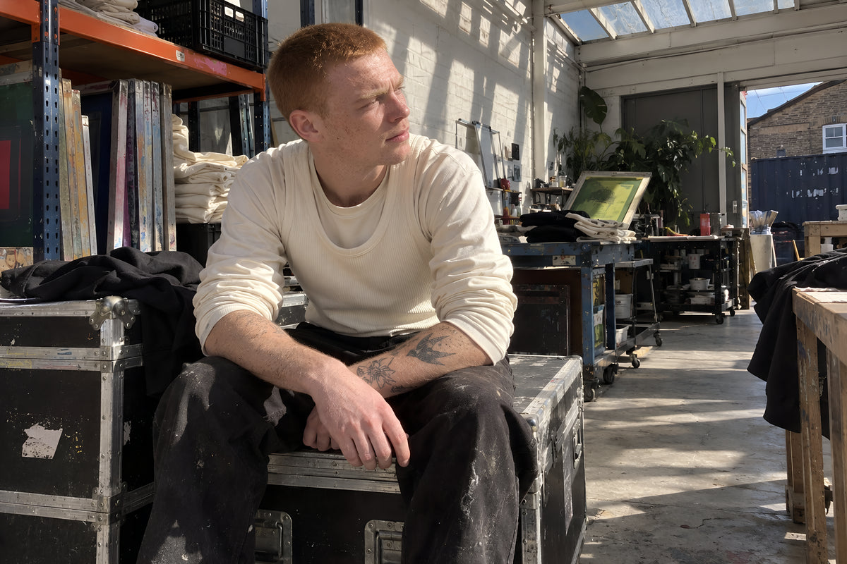 A man with close-cropped ginger hair and freckles sitting on a stack of flight cases in the print room, wearing the bone ENVY Tank over a pushed-up ecru Longsleeve, forearms on knees, looking at something off to the side. Daytime, hard sun through the roof lights, no candles lit.