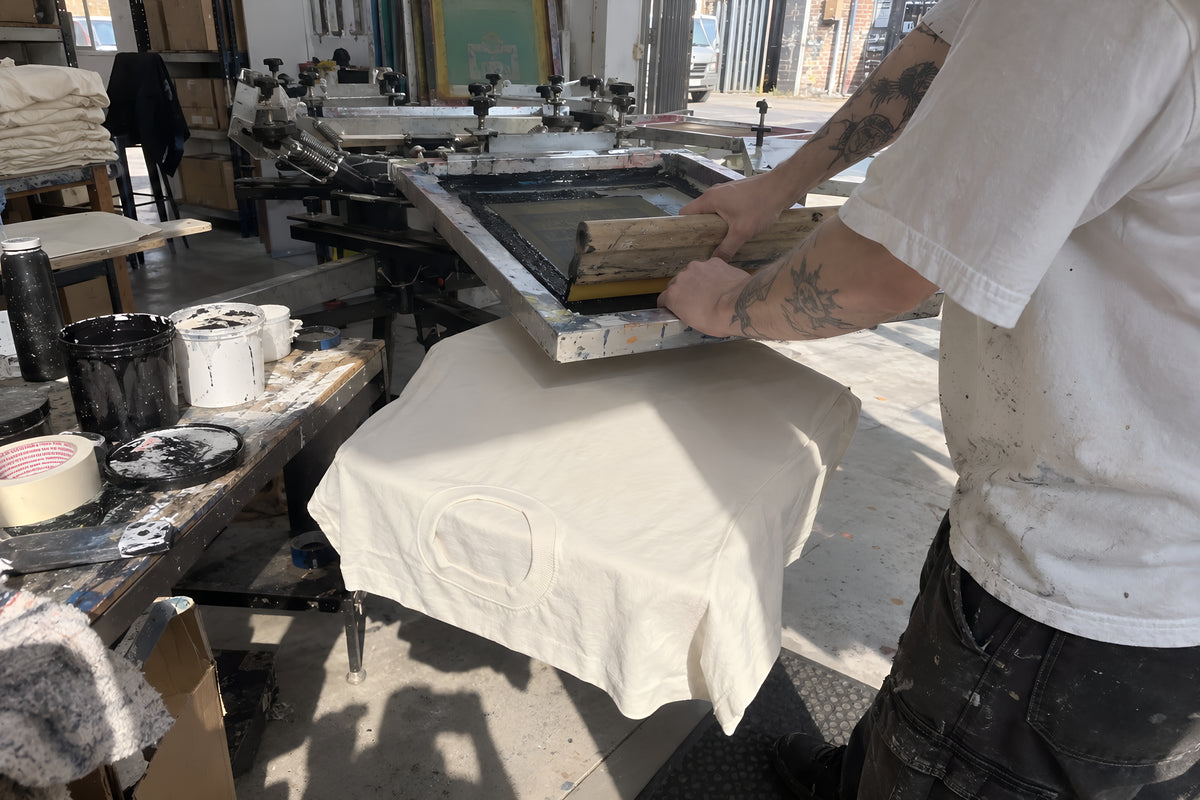 A pair of hands pulling a squeegee down a screen on a carousel press, printing onto a bone ENVY Classic Tee laid on the platen, ink tins and tape all over the bench beside it. Daytime, hard overhead shop light, no candles lit.