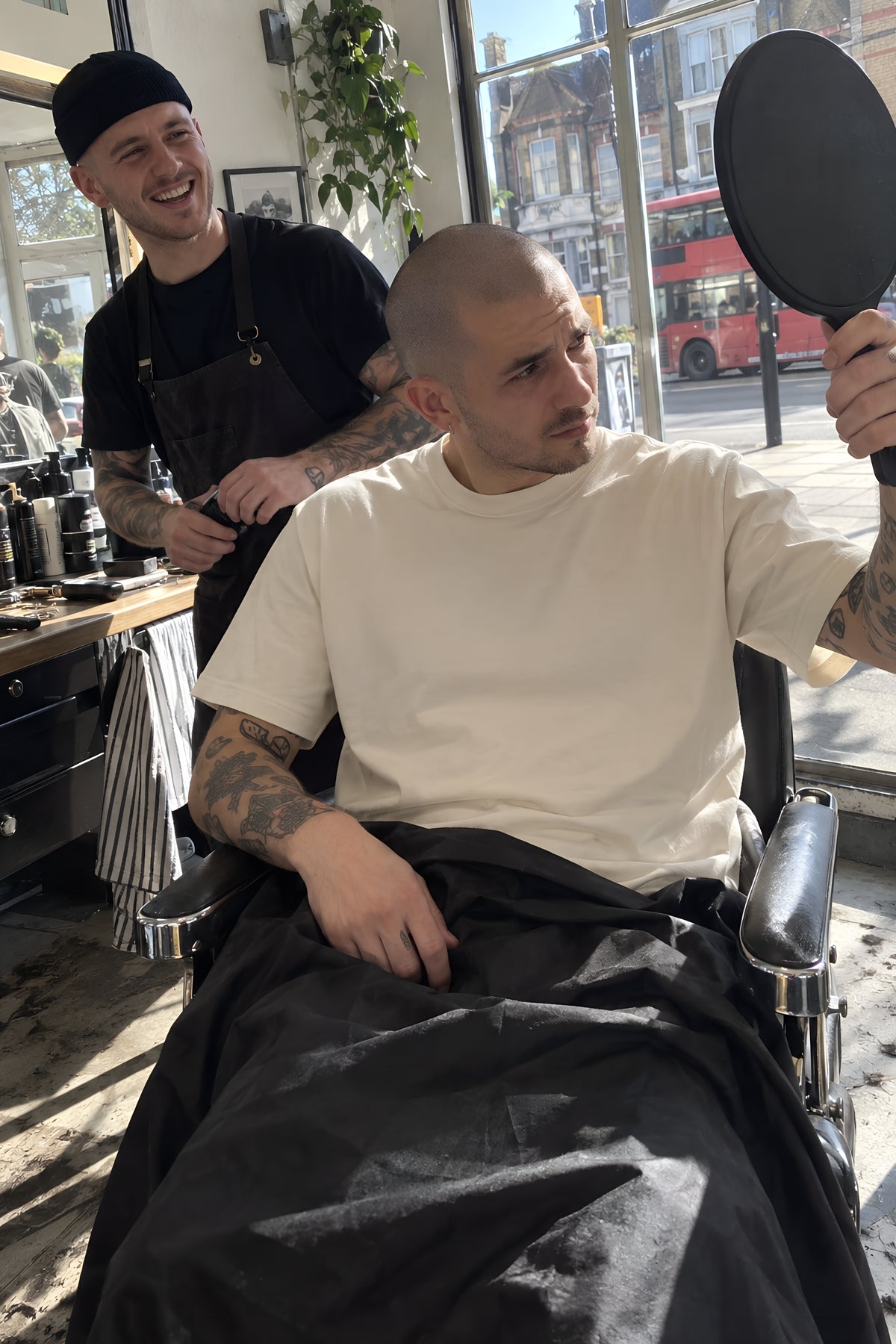 A young man getting a fade in a barber's chair, the black nylon cape pulled down to show the bone ENVY Classic Tee underneath, checking the back with a hand mirror. Daytime, hard sun through the shopfront, no candles lit.