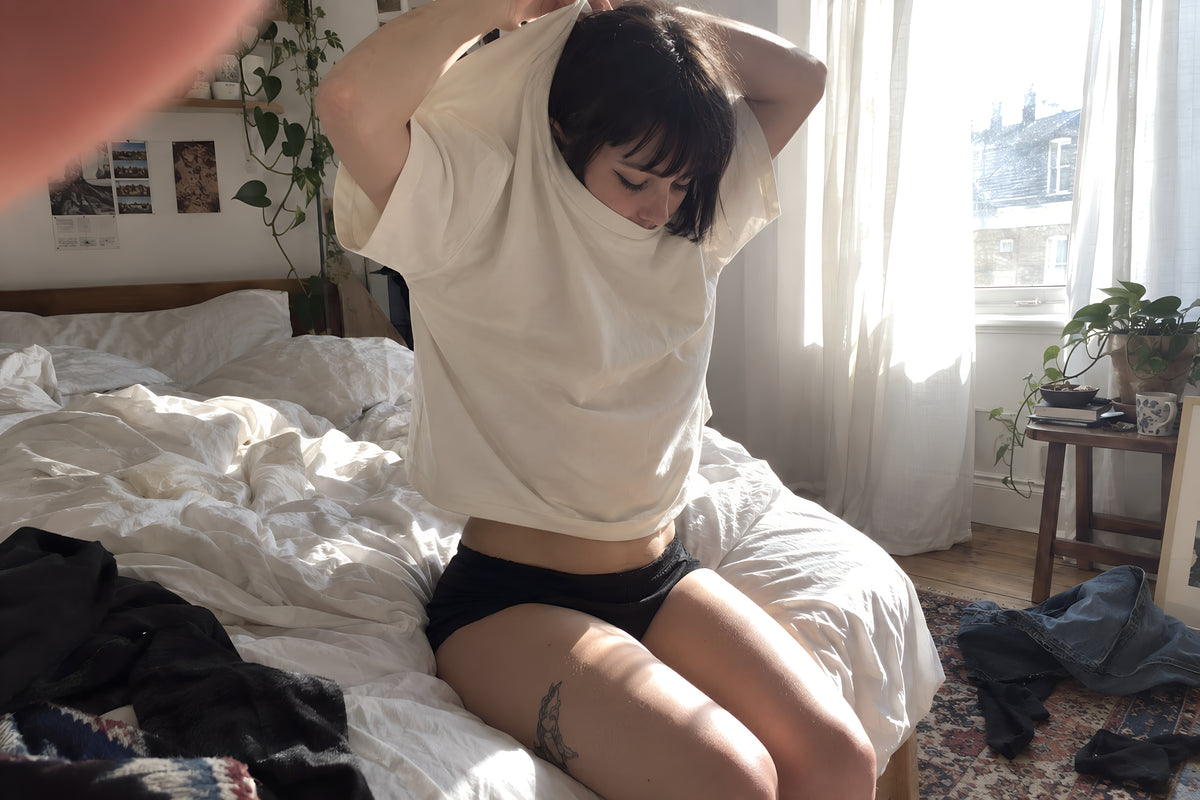 Someone sitting on the edge of an unmade bed pulling the bone ENVY Classic Tee down over their head, duvet shoved back, clothes on the floor. Daytime, hard sun through thin curtains, no candles lit.