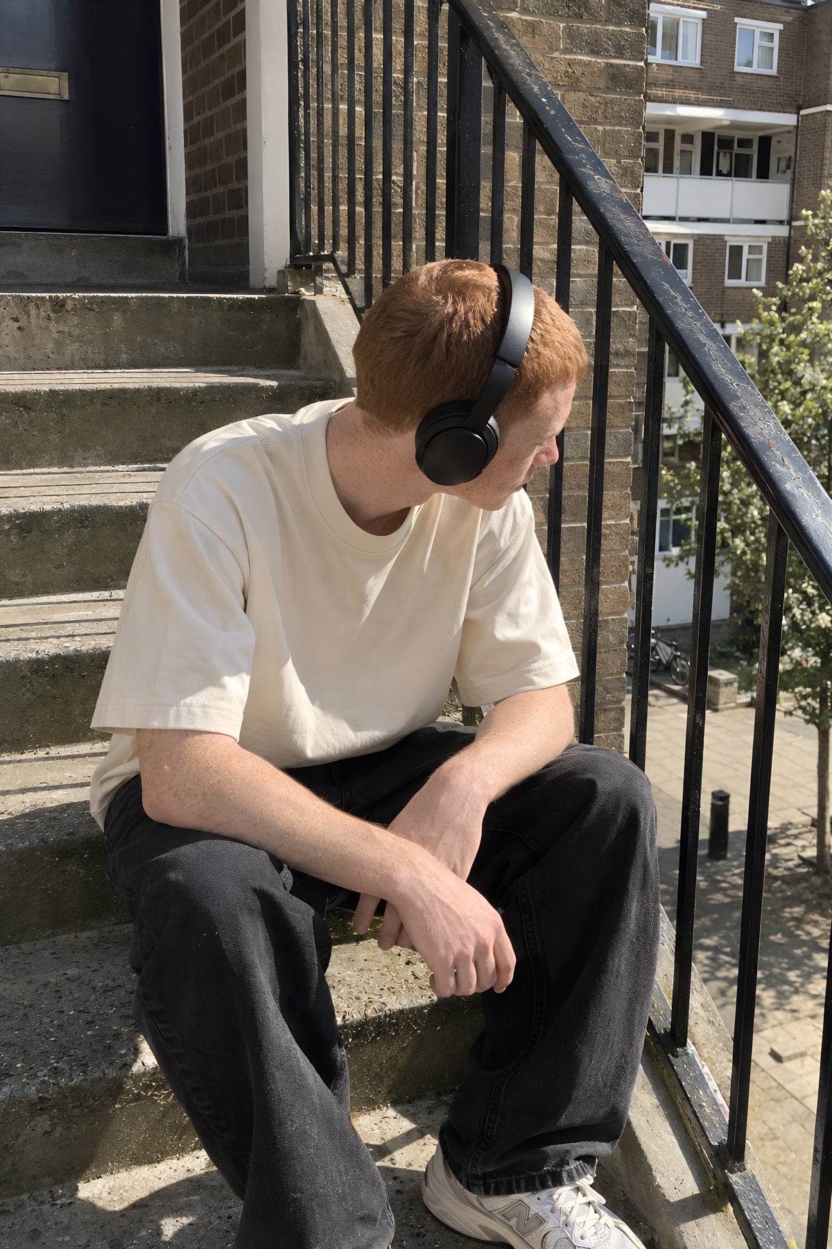 A young man sitting on the concrete stairs of a block of flats in the bone ENVY Oversized Boxy tee, headphones on, looking down the stairwell. Daytime, hard sun, no candles lit.