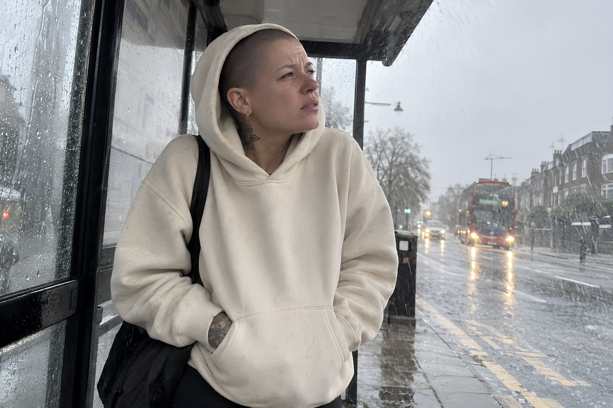 Someone hunched under a bus stop shelter in the sand ENVY Hoodie with the hood up, rain coming in sideways, looking up the road for the bus. Overcast gray daylight, no candles lit.
