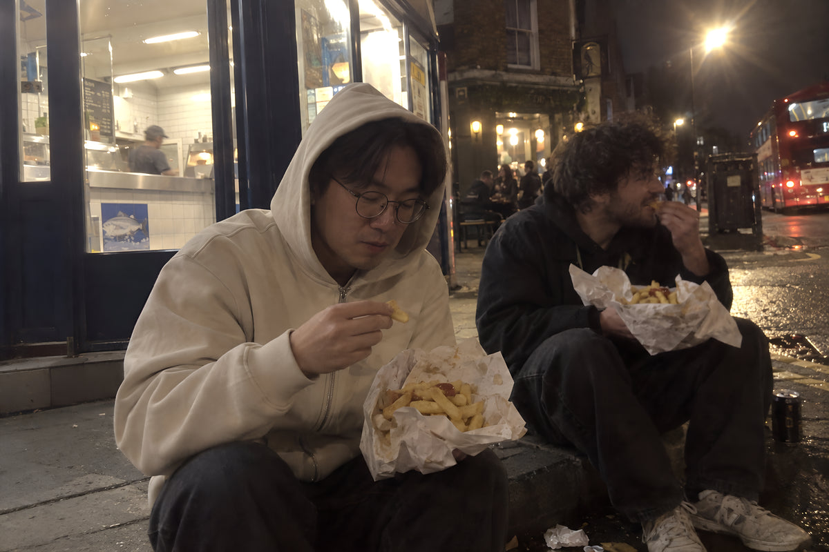 Two people eating chips out of paper on the curb outside a chip shop late at night, one in the ENVY Zip Hoodie with the hood up.
