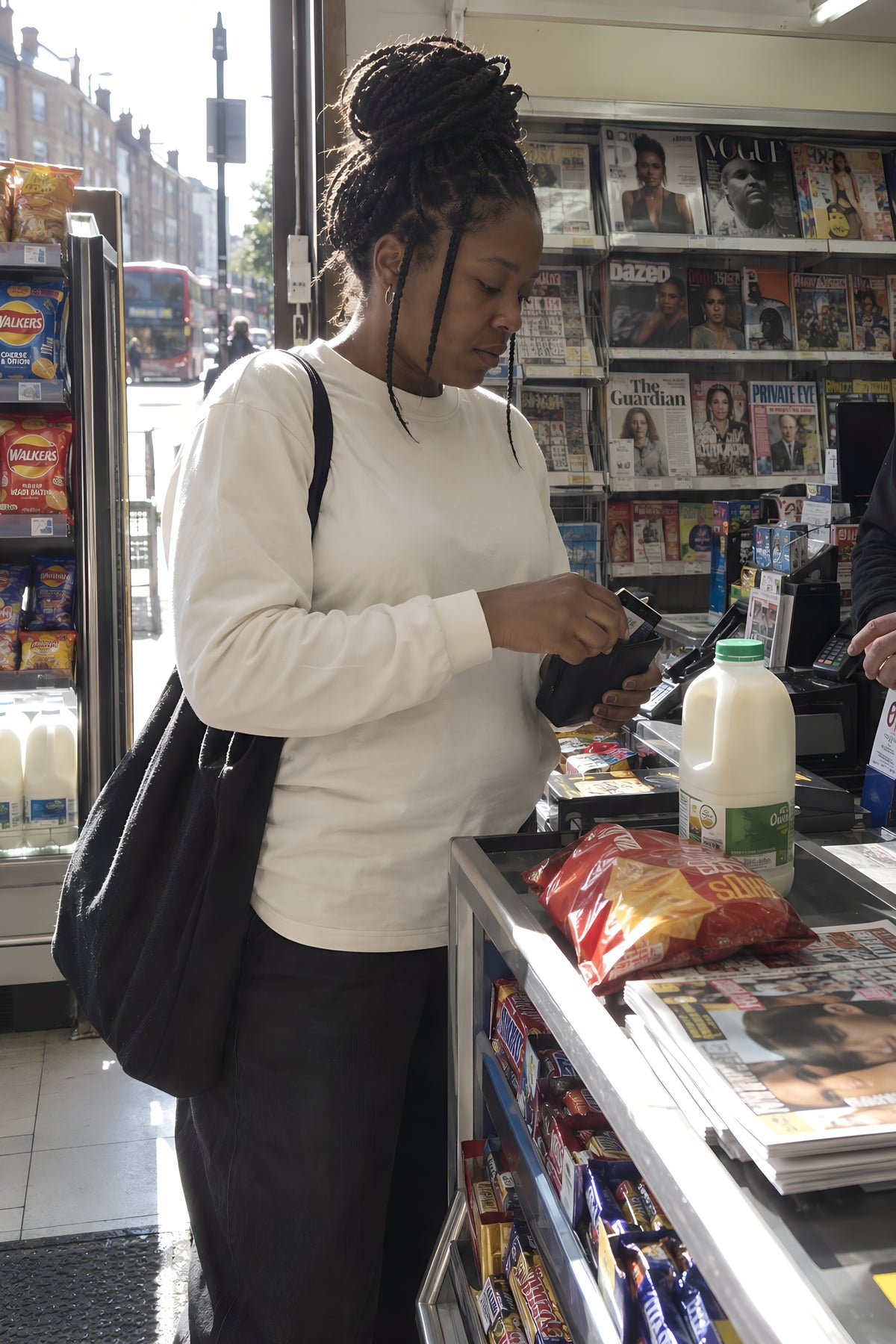A woman in the ecru ENVY Longsleeve at the counter of a corner shop paying for milk, crisps and magazines racked behind her. Daytime, strip light, no candles lit.