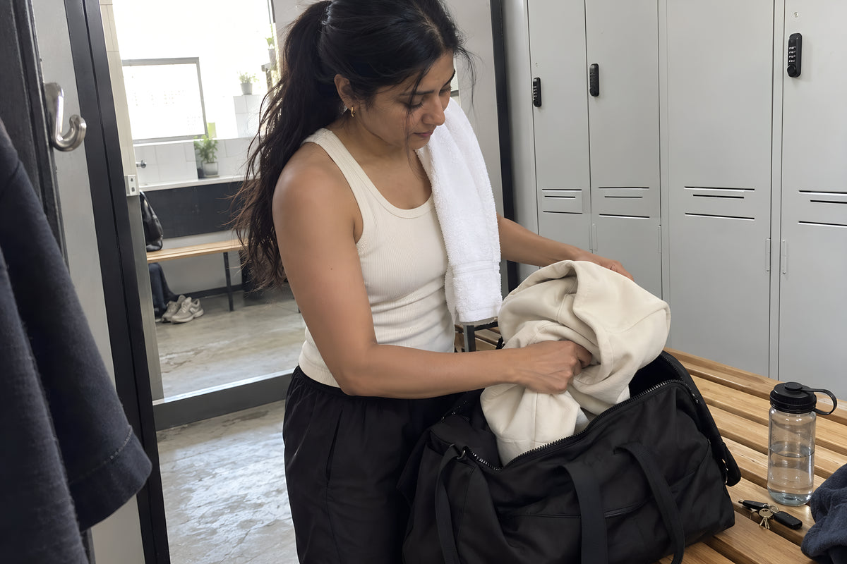 Someone stuffing the sand ENVY Hoodie into a holdall on a changing room bench, the bone Tank already on, towel over the shoulder. Daytime, strip light, no candles lit.
