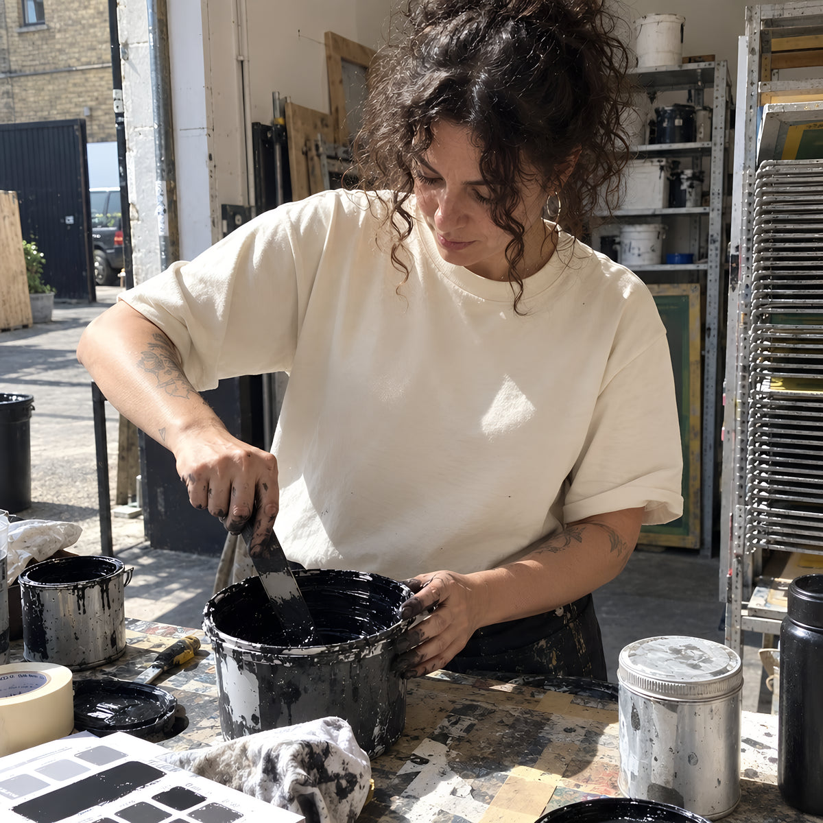 Hands stirring a tub of black ink with a palette knife on a bench covered in tape, tins and color chips, the sleeve of a bone ENVY Classic Tee pushed up. Daytime, hard sun, no candles lit.