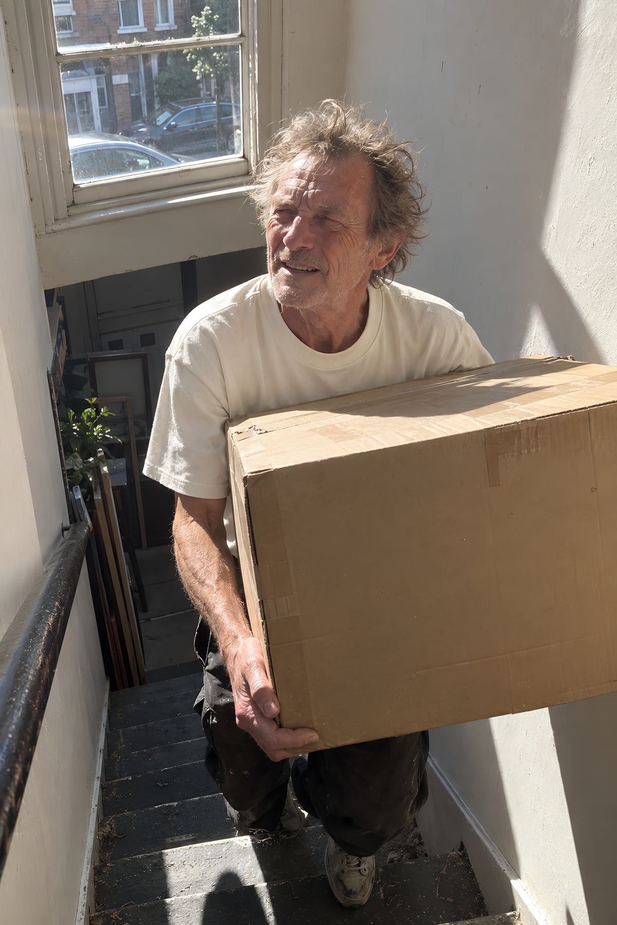 A man carrying a heavy cardboard box up a narrow staircase in the bone ENVY Heavyweight 220 tee, arms braced under it, chin over the top. Daytime, hard sun, no candles lit.