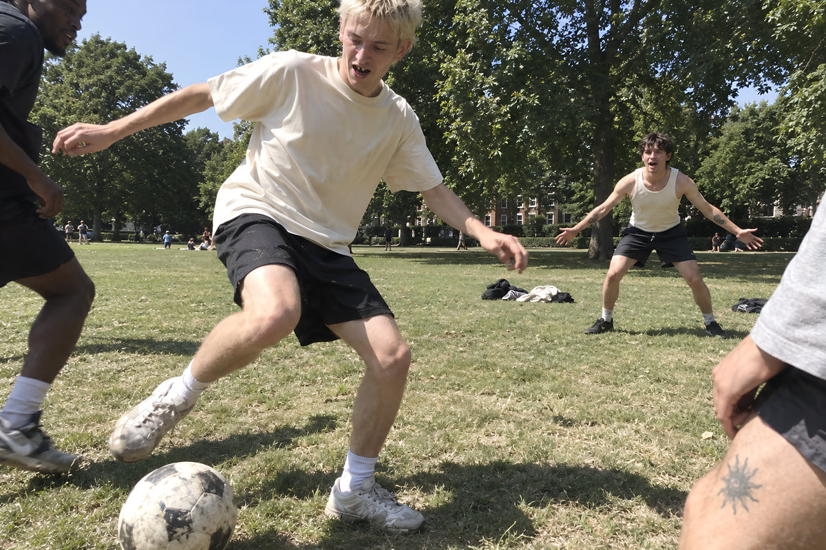 A kickabout in a park with jumpers down for goalposts, someone in a bone ENVY Classic Tee stretching for the ball, another in the Tank standing in goal shouting. Daytime, hard sun, no candles lit.