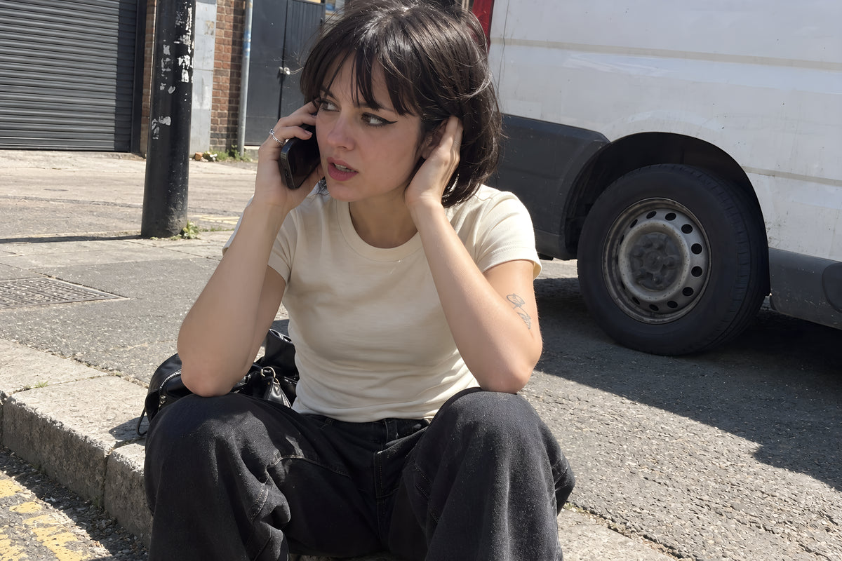 A woman in the bone ENVY Fitted Tee sitting on a curb taking a phone call with one hand over her other ear, van wheels behind her. Daytime, hard sun, no candles lit.