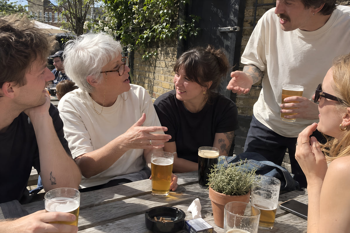 A crowded pub garden bench, five people wedged in with pints, two in ENVY tees, everyone talking across each other and one half standing to make a point. Daytime, hard sun, no candles lit.