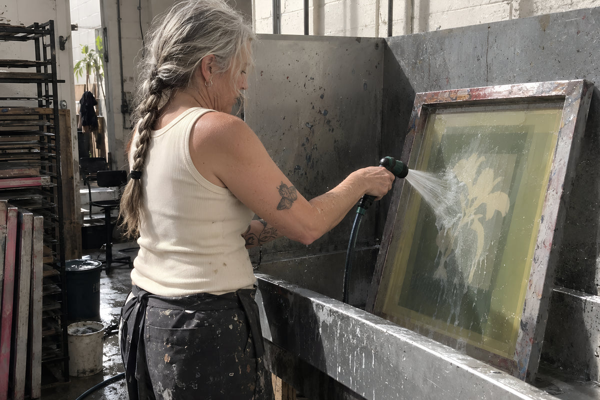 A printer in the bone ENVY Tank hosing emulsion off a screen in the wash-out booth, water everywhere, arms wet to the elbow. Daytime, hard shop light, no candles lit.