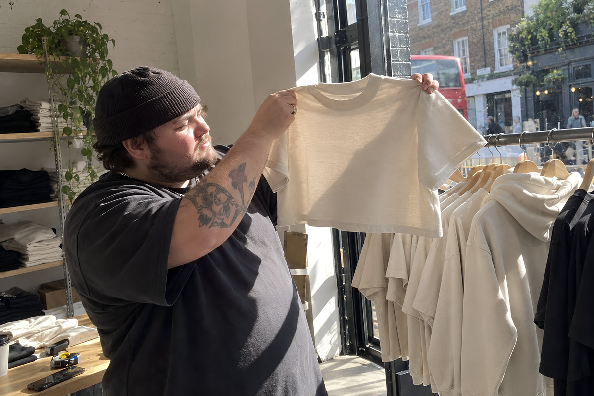 A small shop floor with a rail of ENVY blanks, two friends going through it, one holding a Crop Boxy up to the light to check the weight. Daytime, hard sun through the shopfront, no candles lit.