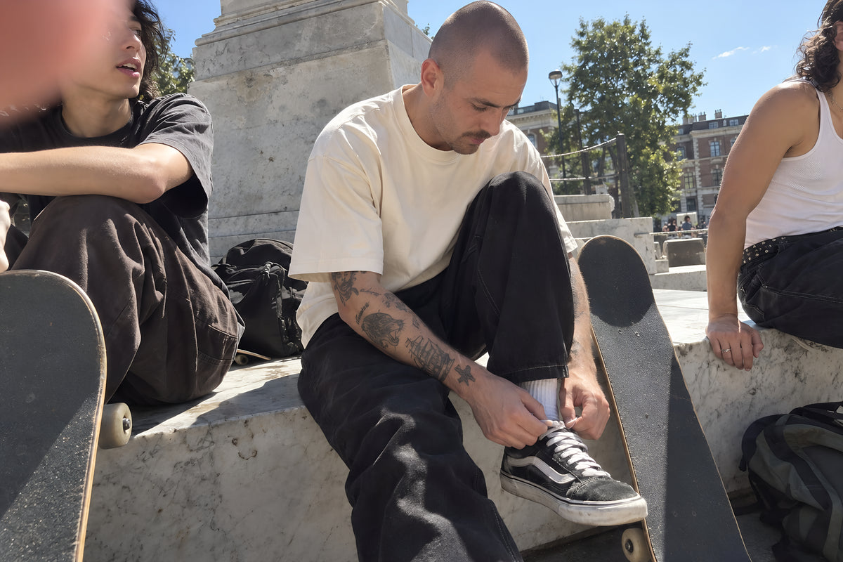 Three skaters sitting along a marble ledge at a skate spot, one in the bone ENVY Oversized Boxy tee re-tying a lace, boards leaning against their shins. Daytime, hard sun, no candles lit.