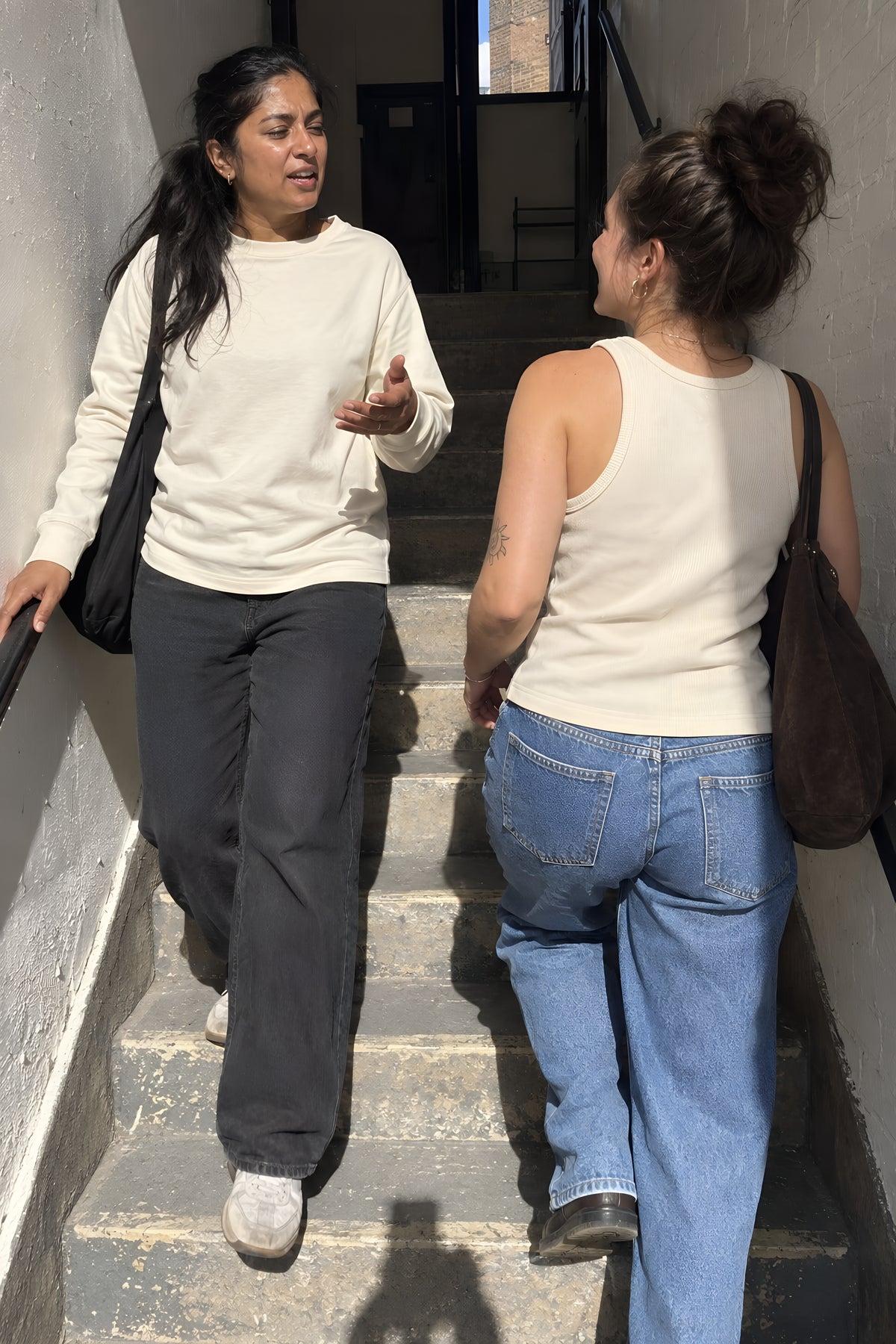 Two women passing on a narrow stairwell, one going up in the bone ENVY Tank and one coming down in the ecru Longsleeve, both half turned mid-word. Daytime, hard sun, no candles lit.