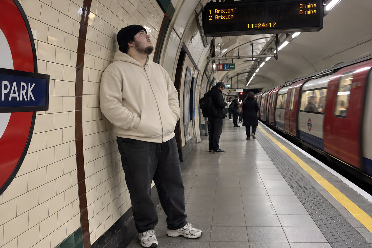 A man in the ENVY Zip Hoodie standing well back from the platform edge on a tube platform, behind the yellow line with his back against the curved tiled wall, looking up at the departure board. He is nowhere near the track edge. Underground, no candles lit.