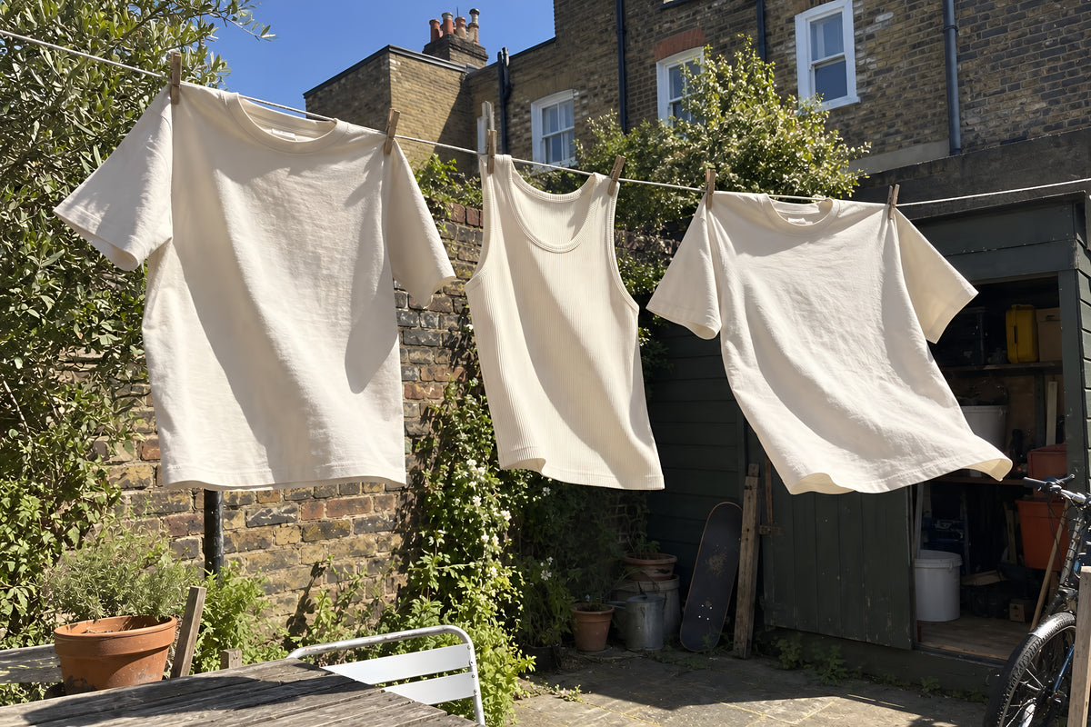 ENVY tees and a tank pegged out on a washing line in a small back garden, blowing sideways in the wind, a brick wall and a shed behind. Daytime, hard sun, no candles lit.